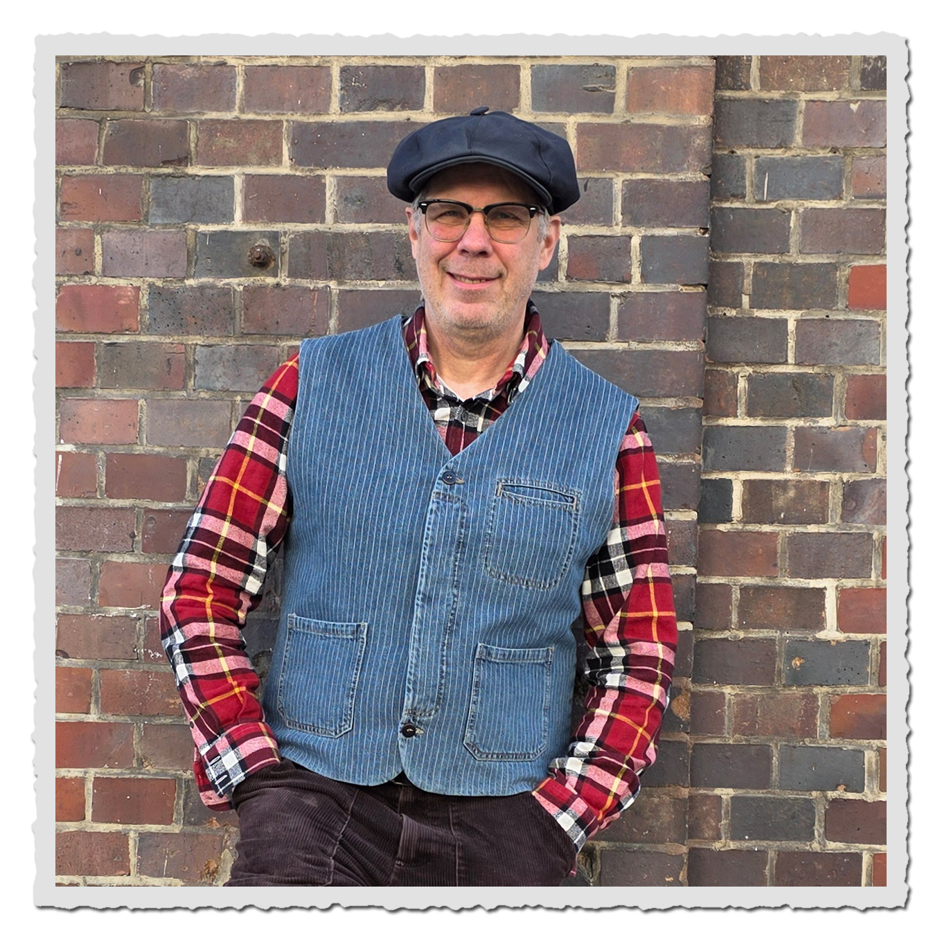 Mann mit Schiebermütze, Brille, kariertem Hemd und blauer Weste vor einer Backsteinwand.